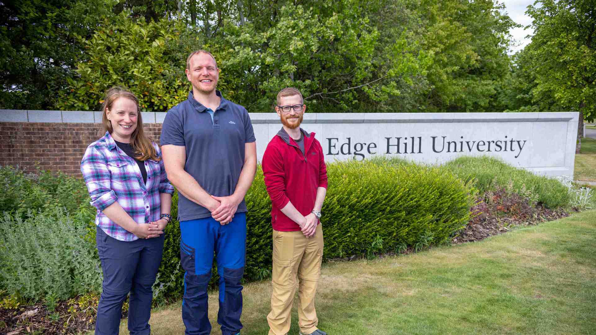 1 female wearing a checked shirt, standing next to a male colleague wearing blue short and he is standing next to another make colleague with a red shirt standing in front of a concrete sign that says Edge Hill University