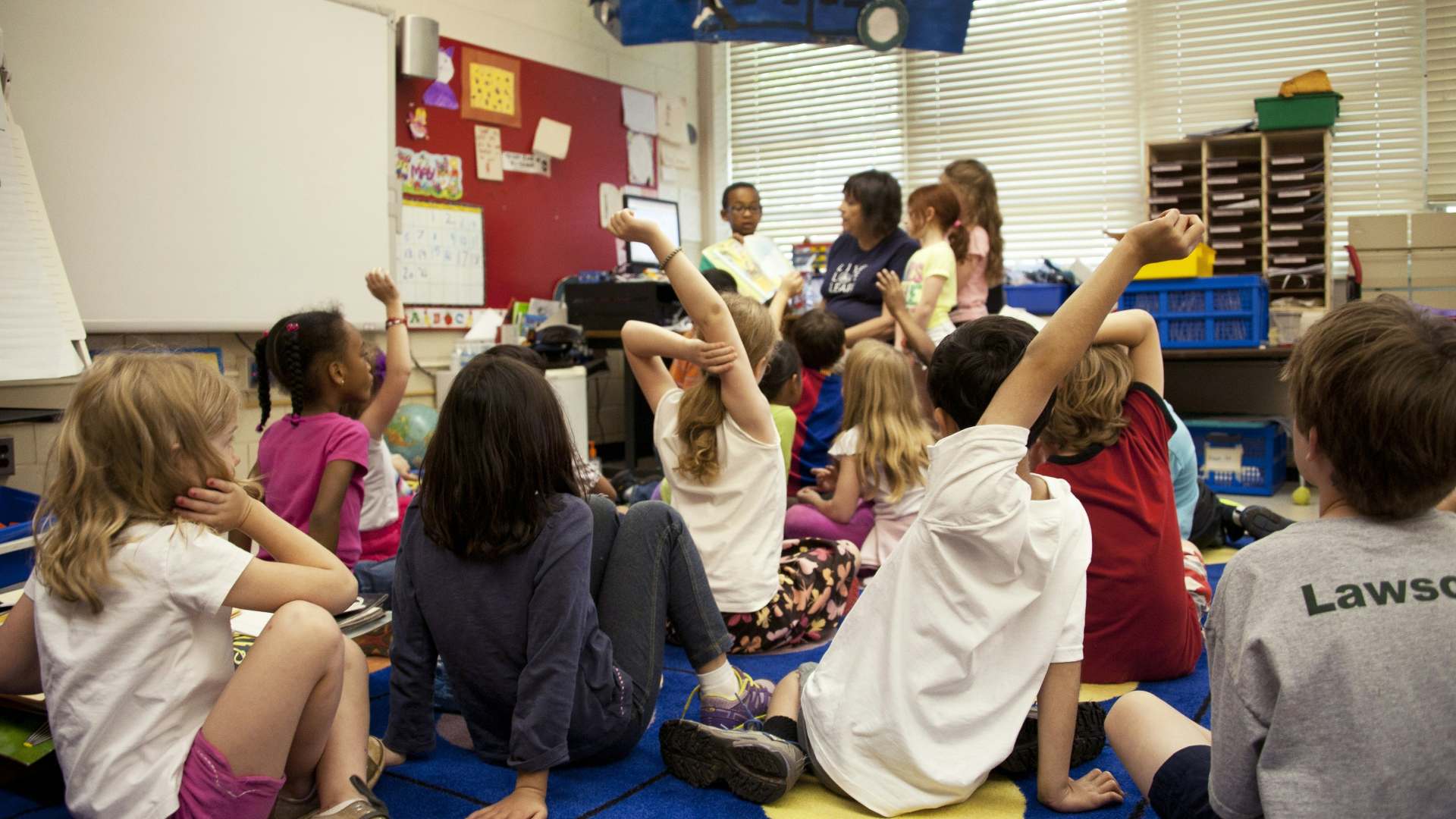 Children sit on classroom floor in front of a teacher, many have their hands in the air.