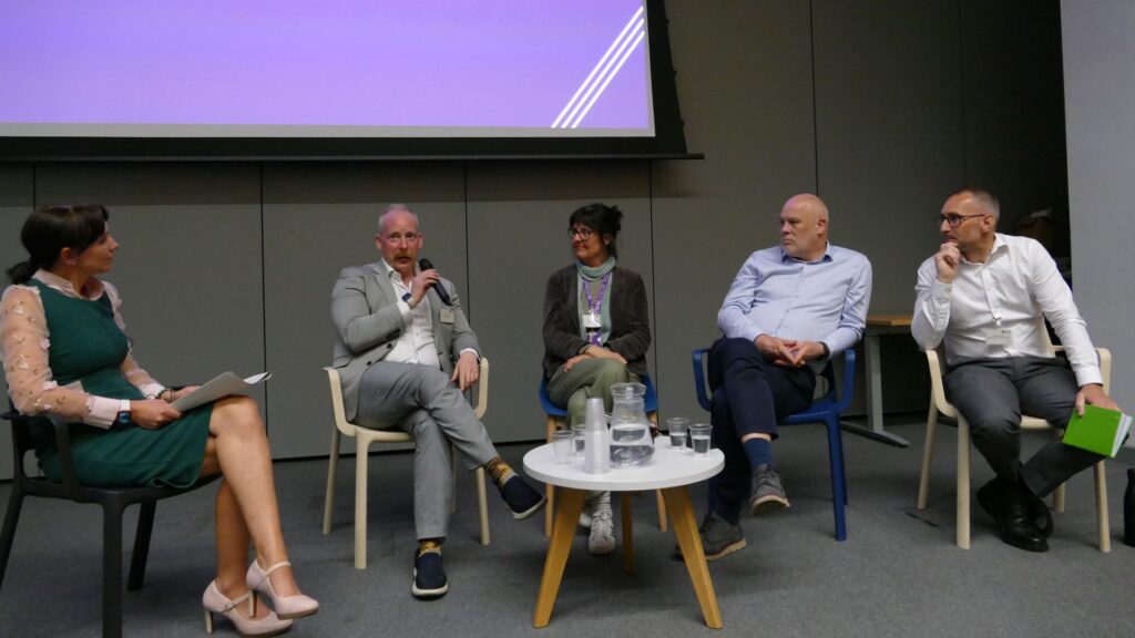 5 panel guest, 2 women and 3 men, all sat down listening to one person holding the mic