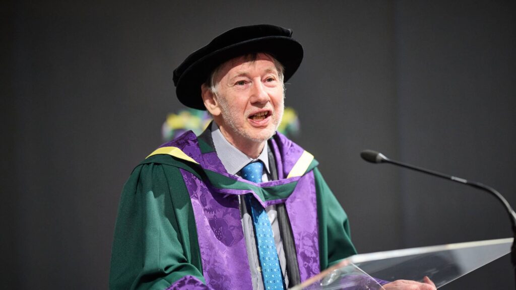 David Sweeney wearing cap and gown stood at a lectern