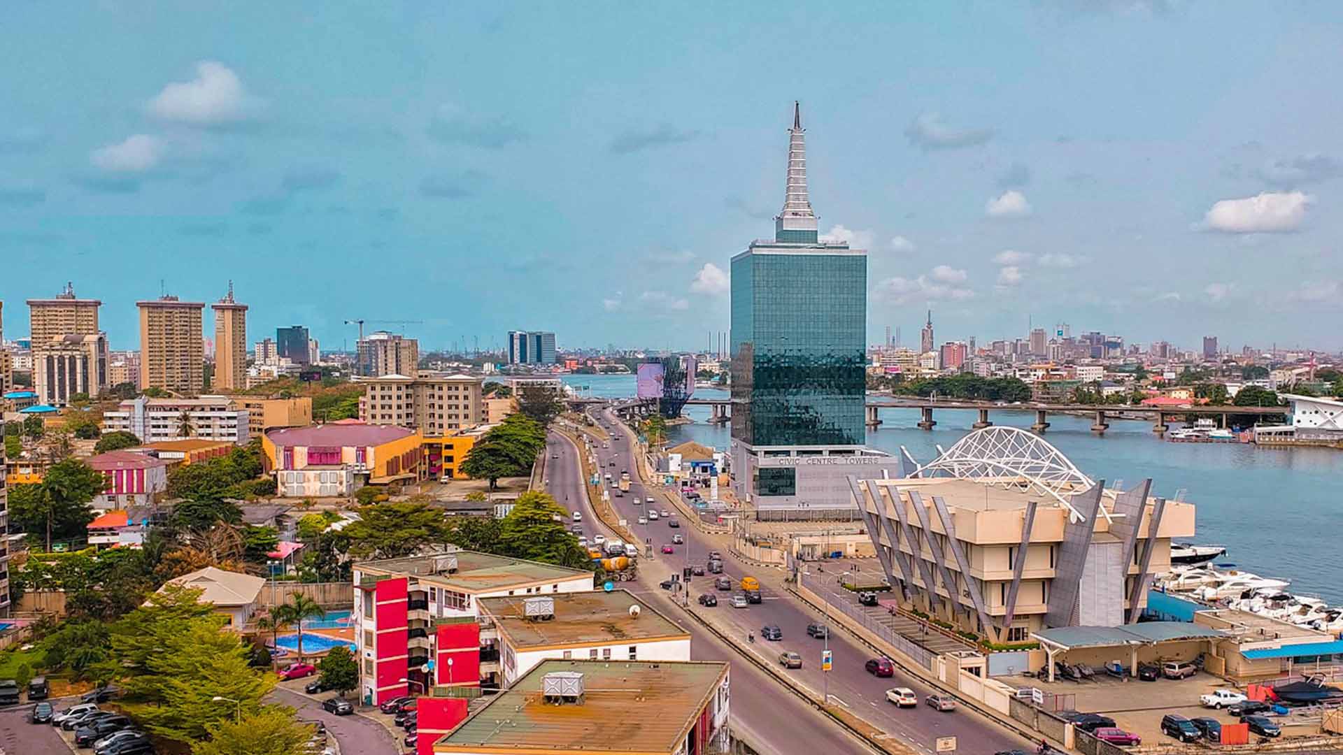 Aerial view of Civic Towers in Lekki, Lagos showing the main road with cars, a lake to the right, a distant bridge, and surrounding buildings.
