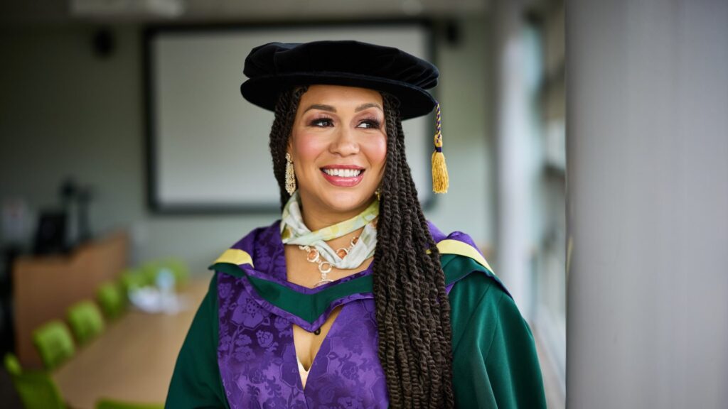 photo of Rebecca Ferguson standing in a classroom with her graduation gown on smiling, looking outside the window.