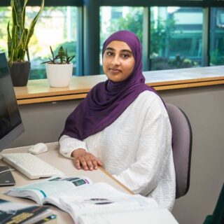 Photo of Esha sat at a computer desk in the Business School. There are books on the table.