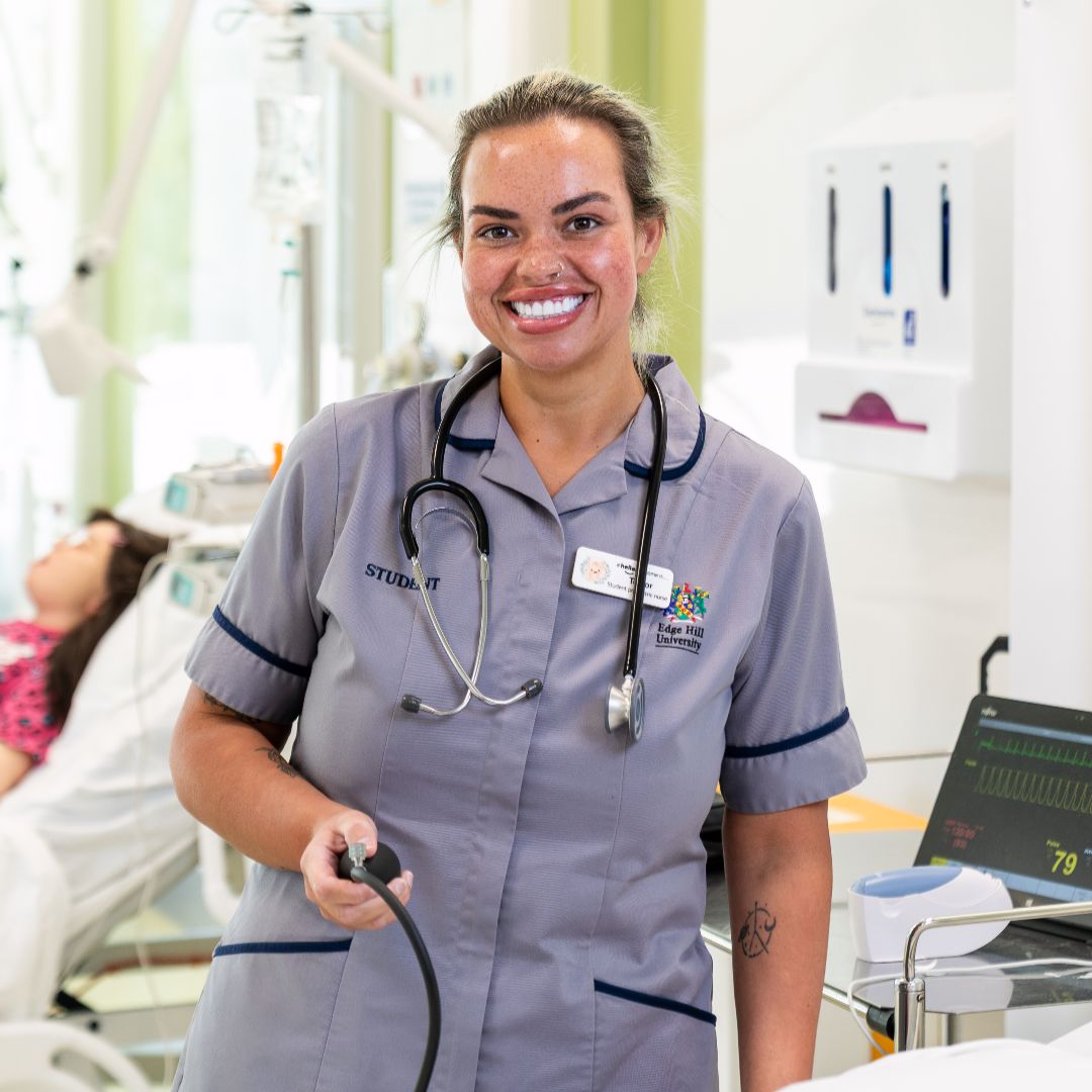 A healthcare student stands in a ward setting with a stethoscope round her neck.