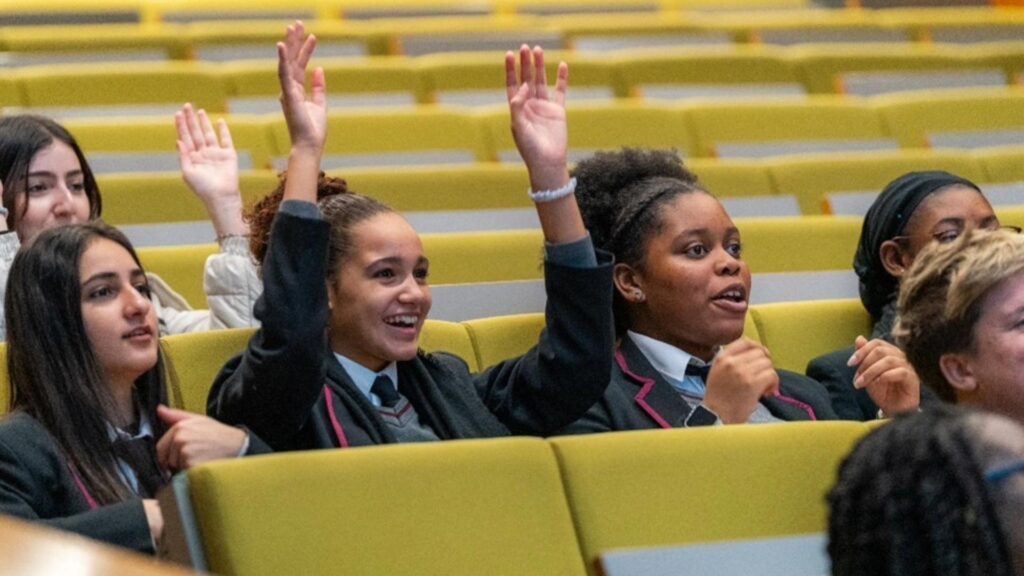 A group of Black, Asian and Minority Ethnic students sitting in a lecture theatre engaging in a presentation by raising their hands.