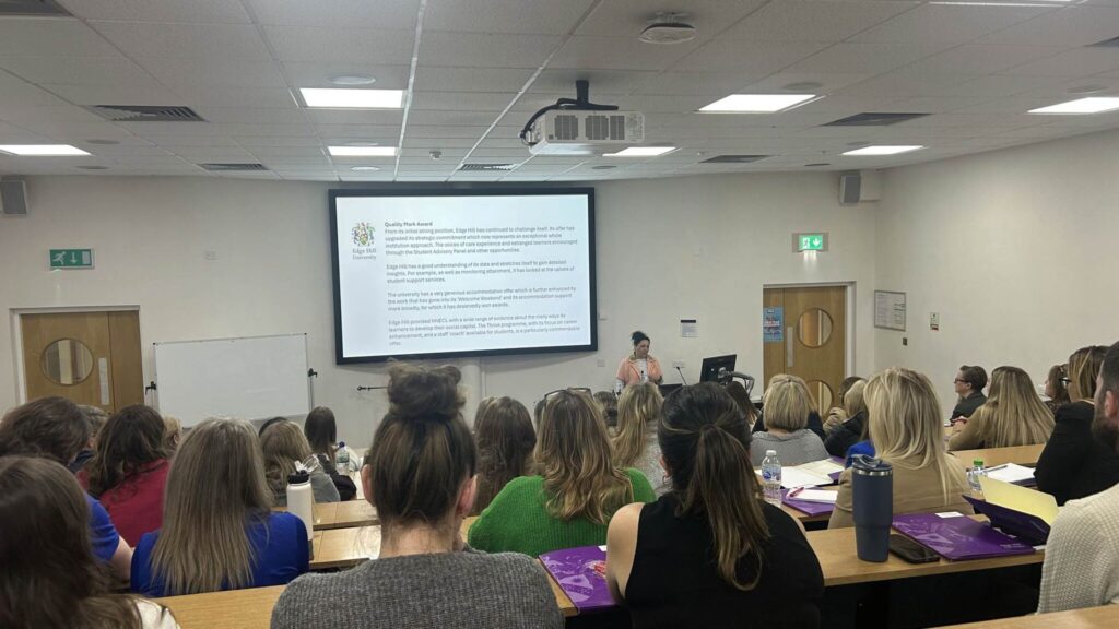 A female speaker is delivering a presentation in a lecture theatre to a full room of attendees who are listening to her speak.