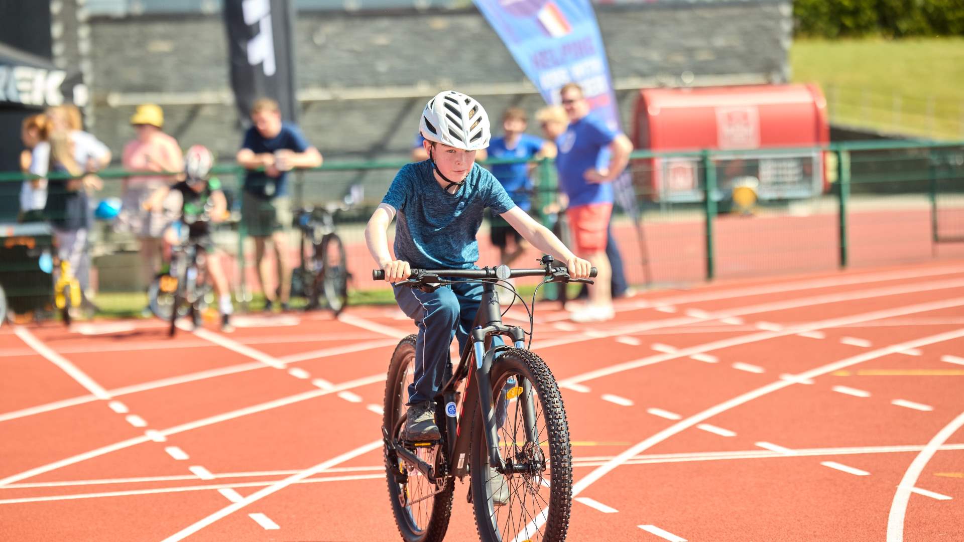 Young boy ridding a bike on a running track and can see other children in the background waiting to ride off.