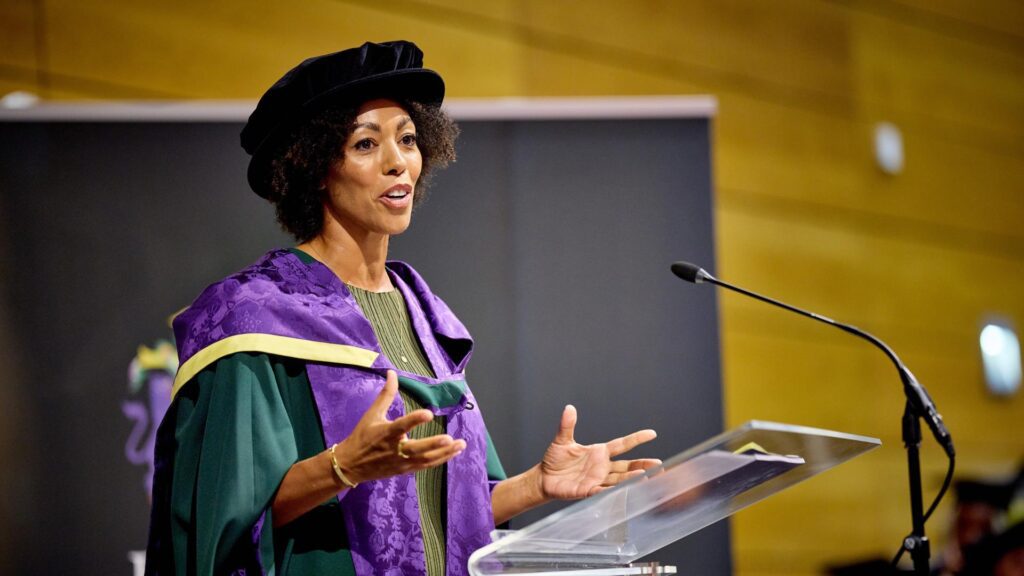 Dr Zoe Williams wears a cap and gown and stands behind a lectern.