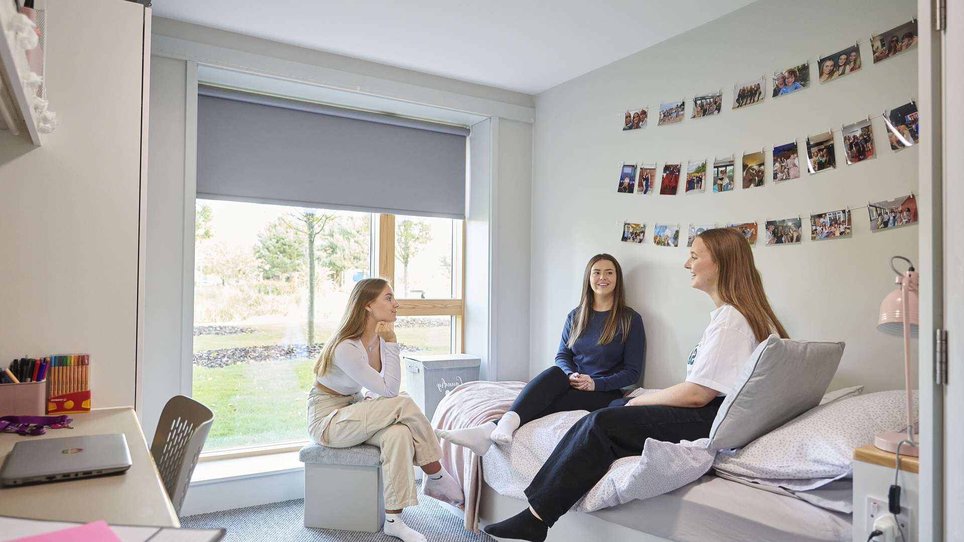 Three students sit in a campus bedroom chatting