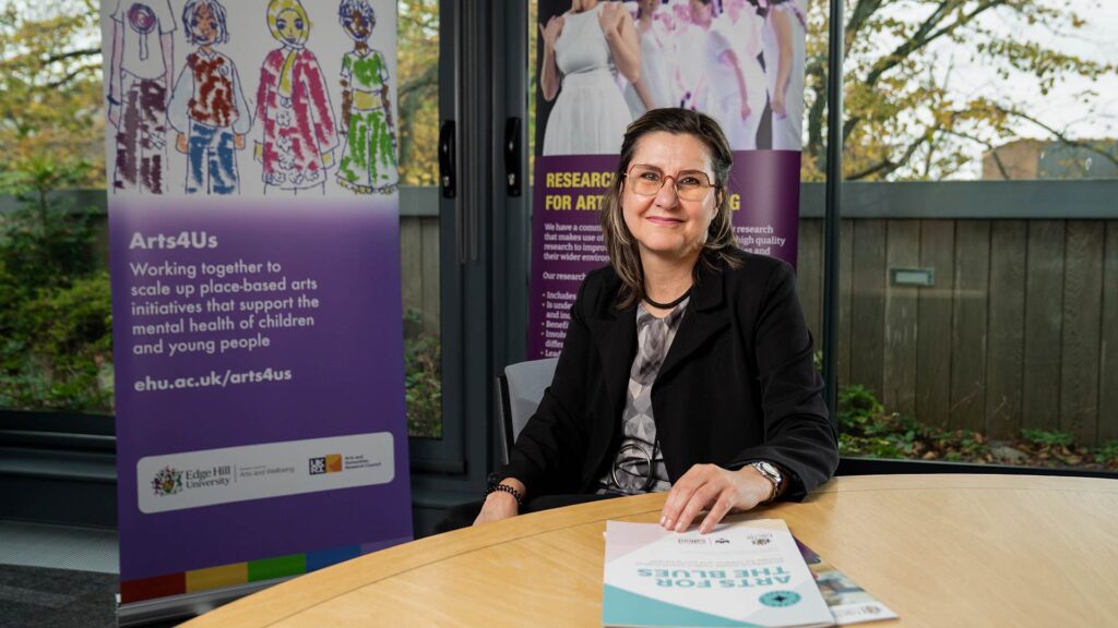 professor Vicky Karkou siting down with two arts for us banner behind her and some flyers on the table in front of her.
