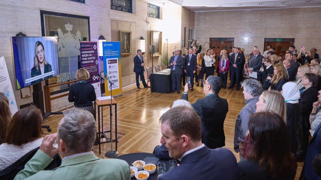 A room full of standing people watch a woman speak from a TV screen.