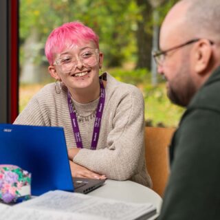 Milo Smith profile photo, sat at a desk with a laptop talking to another student