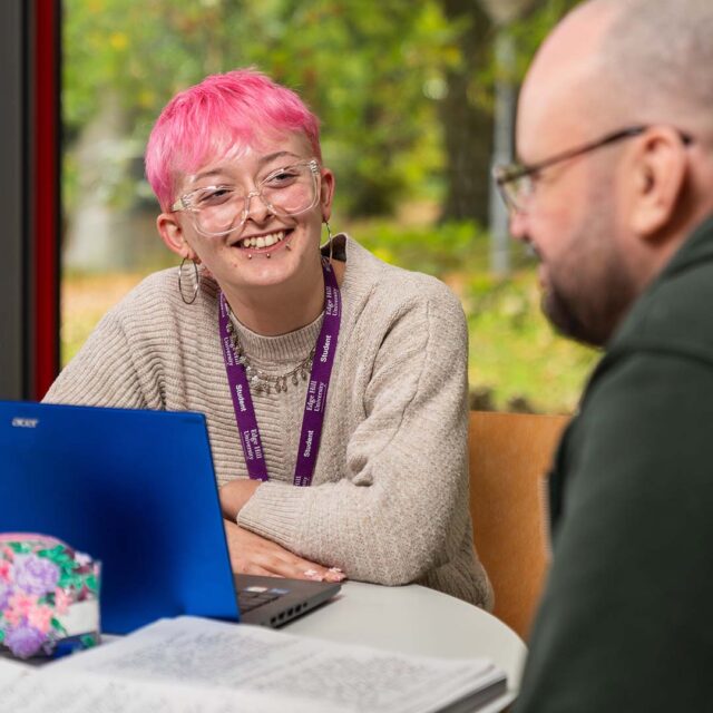 Milo Smith profile photo, sat at a desk with a laptop talking to another student