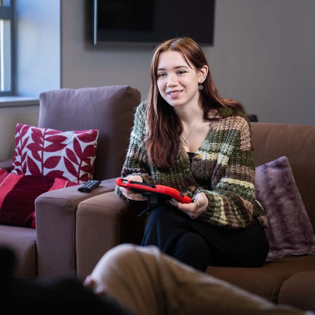 Social Work student sat on a sofa with an electronic device talking to another person