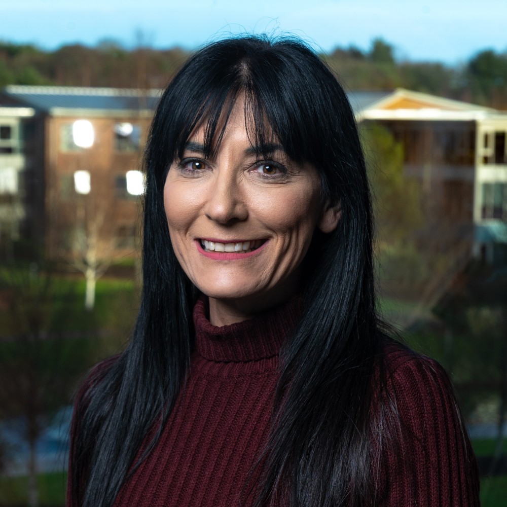 portrait of Abigail standing in front of a window looking strait at camera smiling, wearing a maroon turtleneck jumper. 
