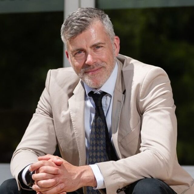 Professor Kurt Allman sits on steps, leans his arms on his knees and smiles the camera.