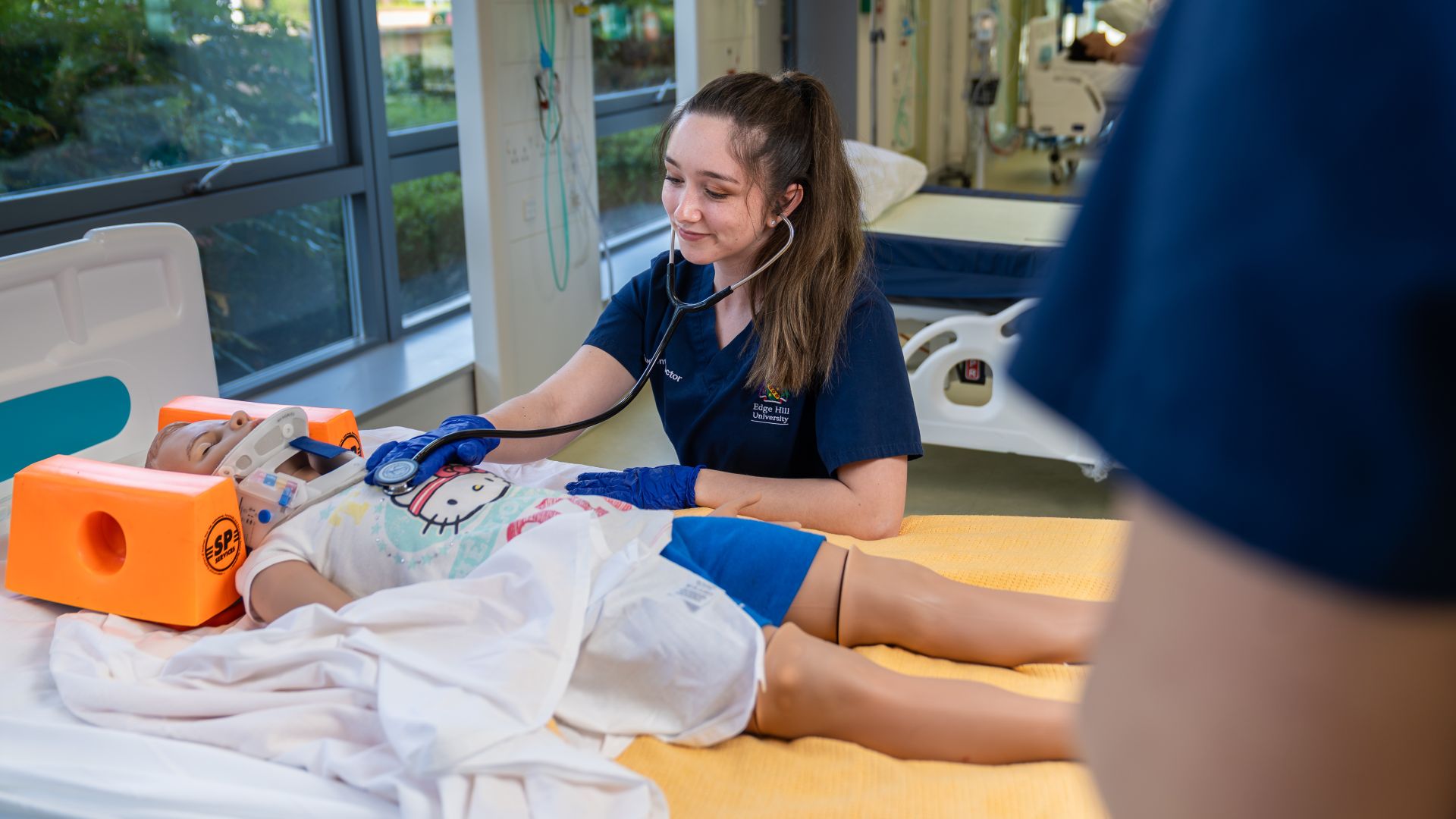 A medical student smiles as she uses a stethoscope to practice listening to a dummy patient’s heartbeat on the ward within the Clinical Skills and Simulation Centre