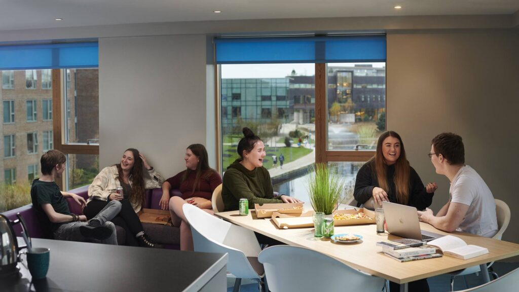 Six students sit socialising on campus in student accommodation while eating pizza.