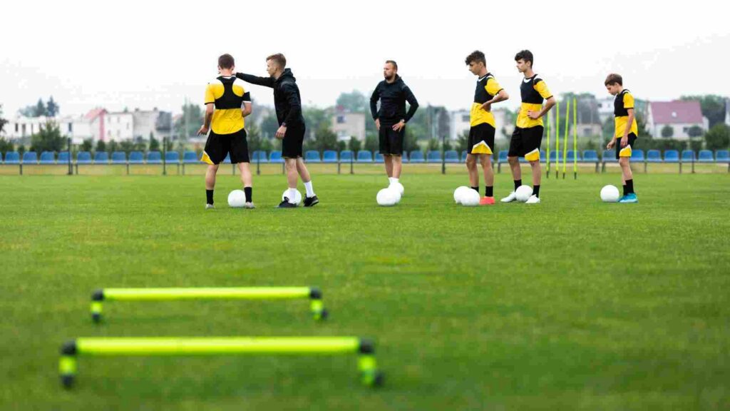 A group of teenage boys and their football coaches stand on a field. There are footballs and equipment around them. 