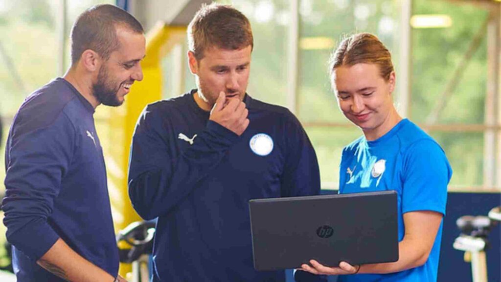Three people stand in a gym. They are looking at a laptop. One is dressed in Wigan Athletic FC training clothing. 