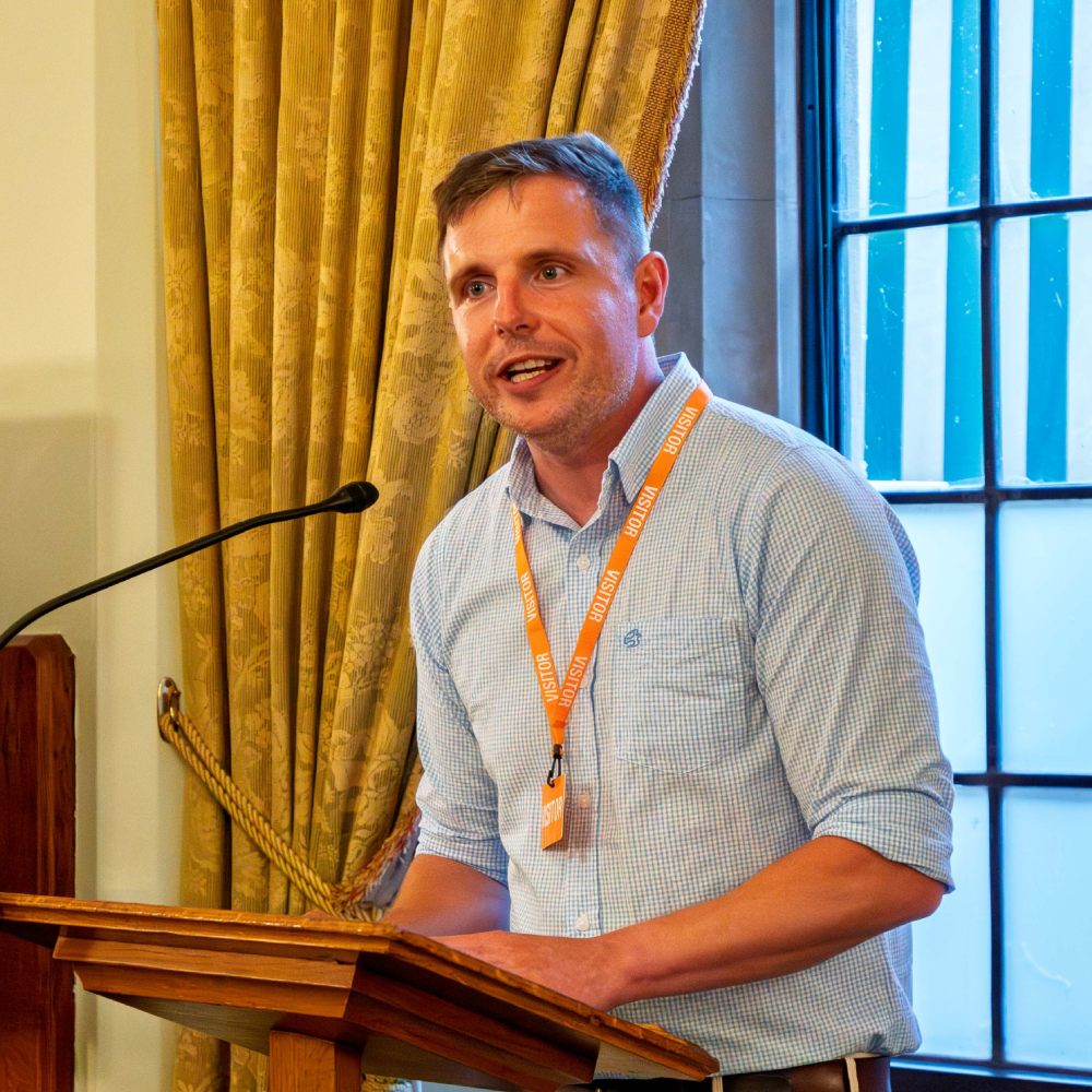 An image of Dr Alex Blower stood at a lectern, speaking into a microphone at one of his professional talks.