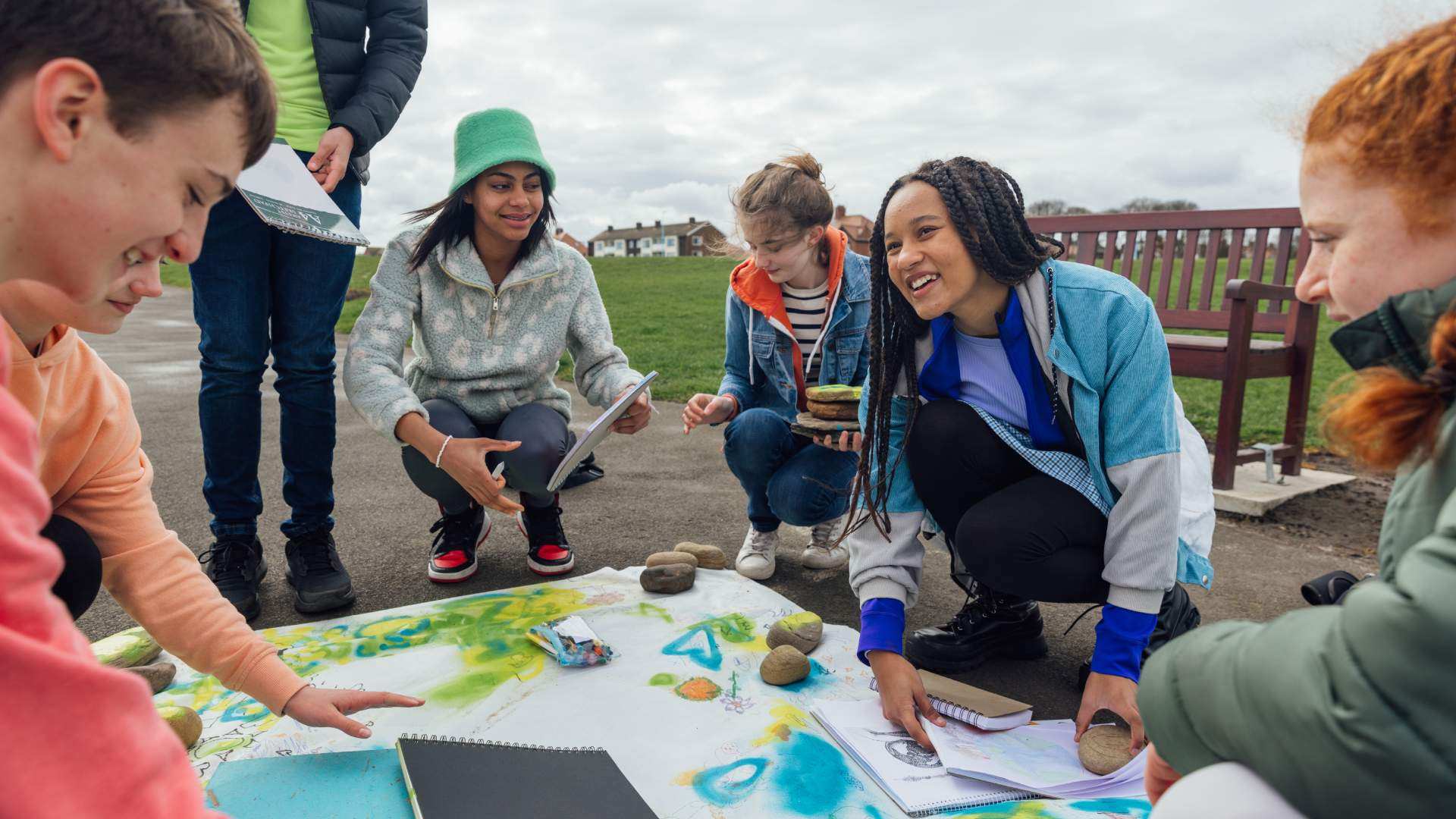 A group of young people kneeling on the ground outside, working on an art project, holding sketch books in their hands.