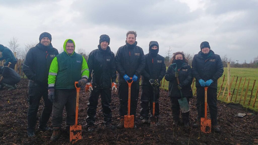 A group of people stand in a line wearing hi vis and holding long-handled spades.