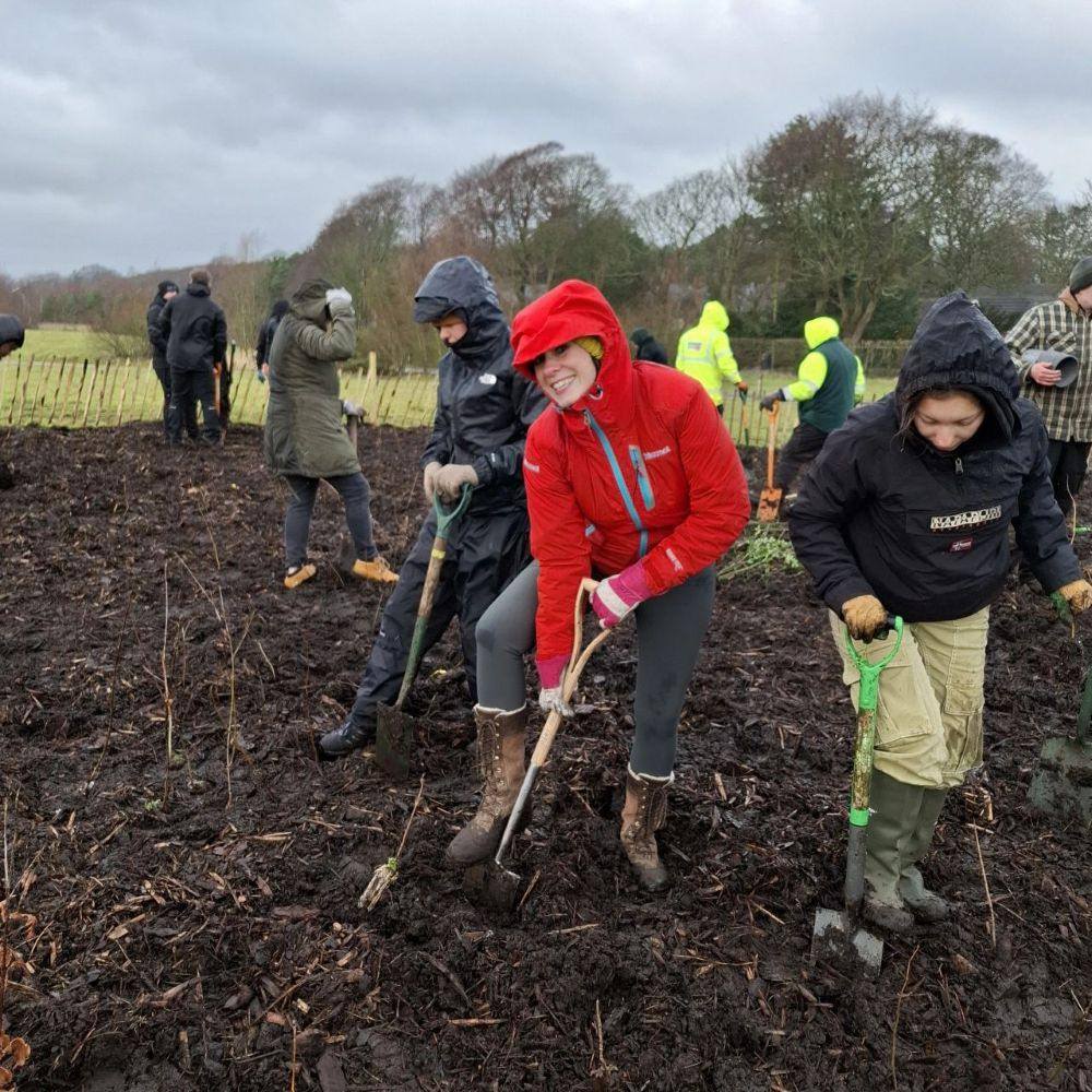 Students wearing wellies and raincoats dig the soil with long-handled spades.