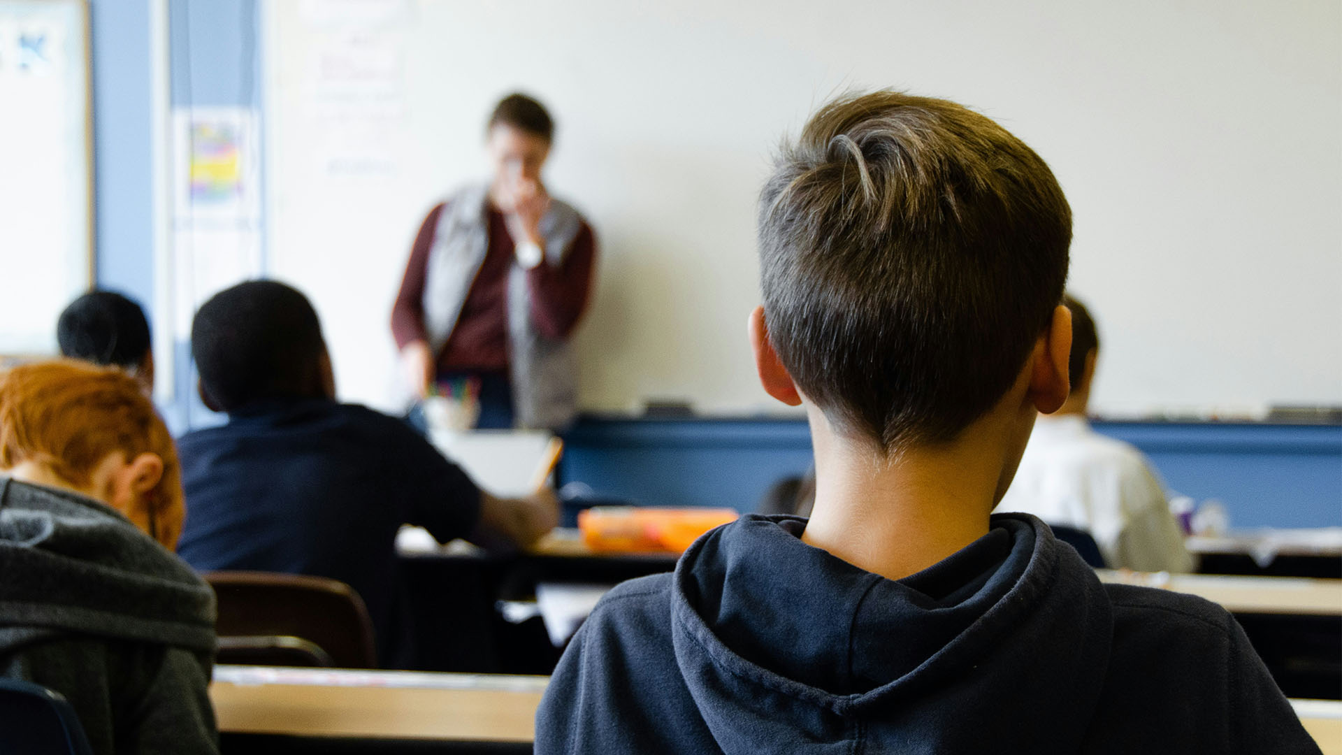 Image of a classroom with a teacher stood at the front of the class and children at their desks.
