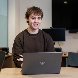 Matthew Wallace working on their laptop, smiling at their friend