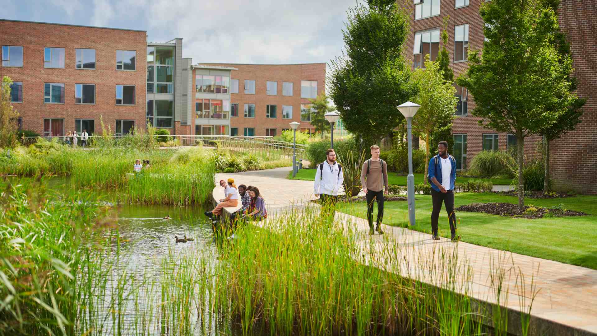 Landscape image of three Edge Hill University students walking on campus past two students sat by a lake and student accommodation.