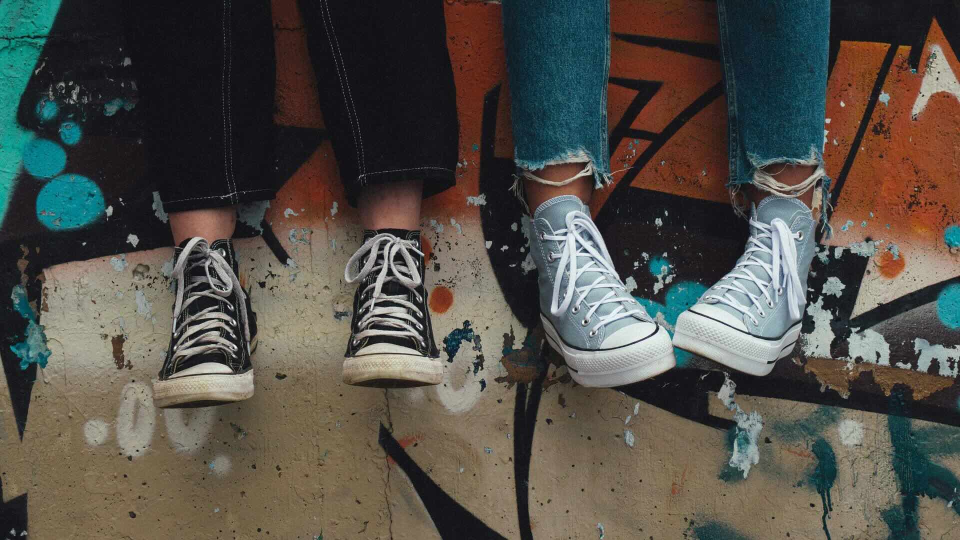 two feet dangeling from a graffiti wall - both people wearing skinny jeans and converses