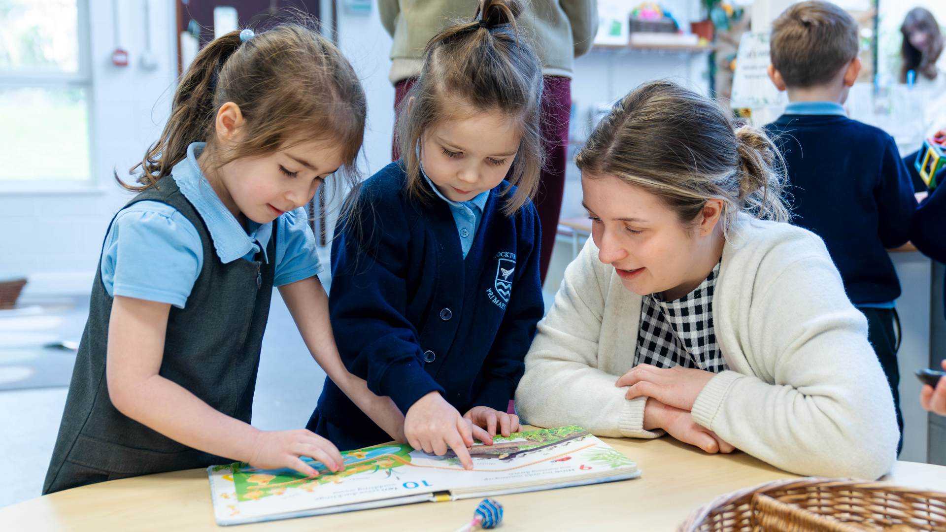 Trainee teacher sat at desk with two primary aged pupils in a classroom setting looking at a book together