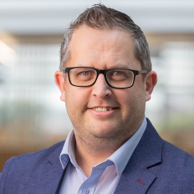 portrait photo of Andy - looking into camera smilling, wearing black glasses and blue shirt with blue suit