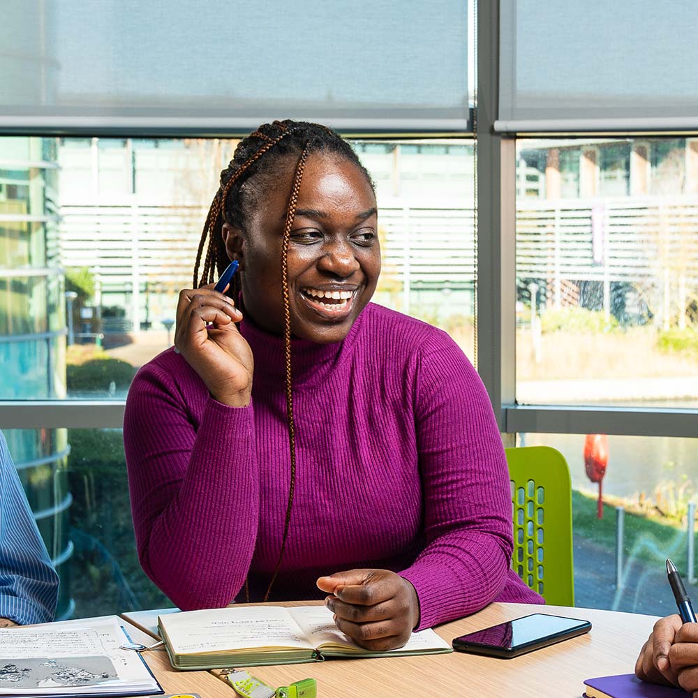 Student Chika sits at a table in a bright study space by large windows, holding a pen while looking to the side. An open notebook, loose papers, a highlighter, and a mobile phone are placed on the table, with part of another person and study materials visible nearby.