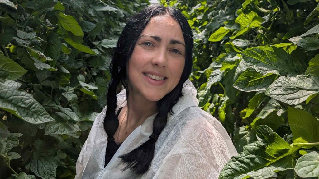A student is in a greenhouse, looking at the camera and smiling.