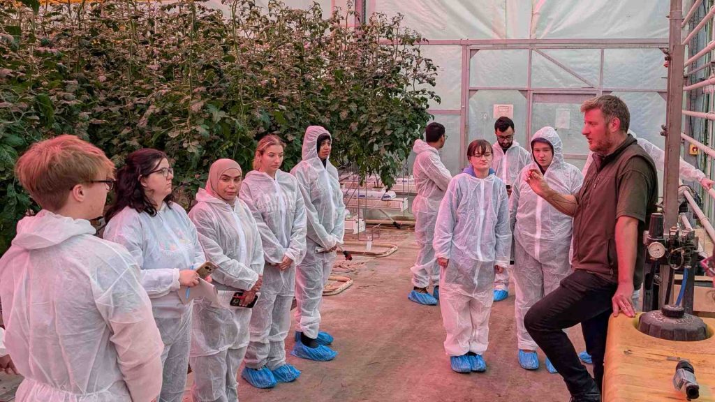 A group of students are in a greenhouse, all wearing hazmat suits. They are having a talk delivered by a man.