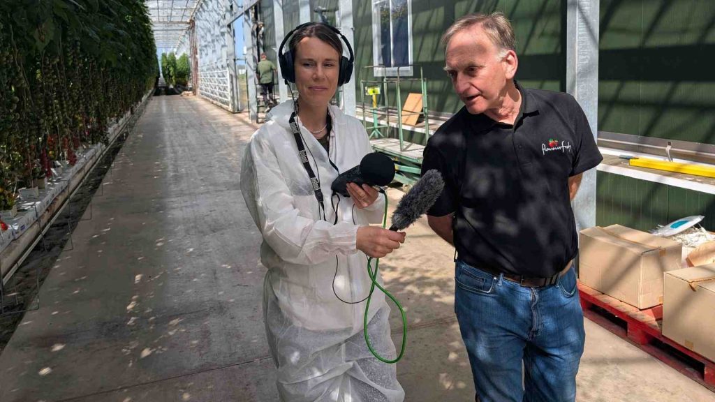A man in a FlavourFresh t-shirt is talking to a woman wearing a hazmat suit. The woman is holding a microphone.