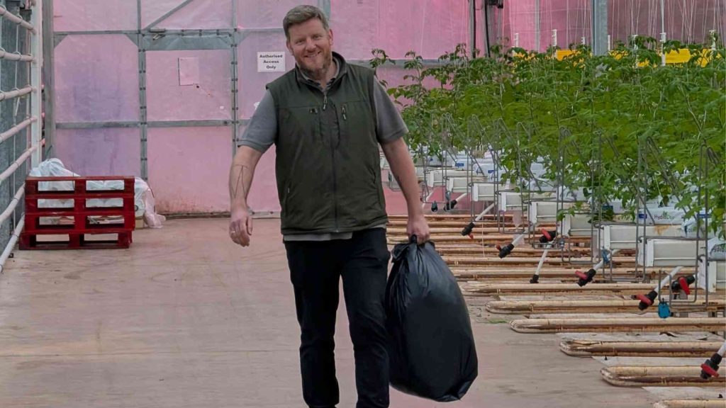 A man in a greenhouse carrying a plastic bag. He is smiling.