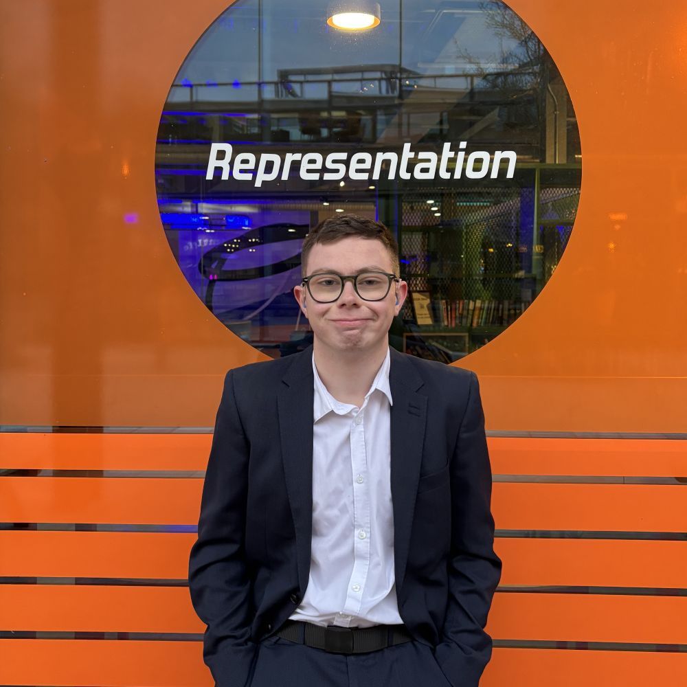 student Ruben standing in front of glass window with orange design and the word representation above him. Ruben is wearing a white shirt and dark blue suit.