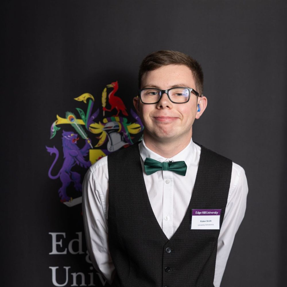 student Ruben Smith wearing glasses a green bow tie with a black waist coat and white shirt, standing behind banner with Edge Hill crest on it.
