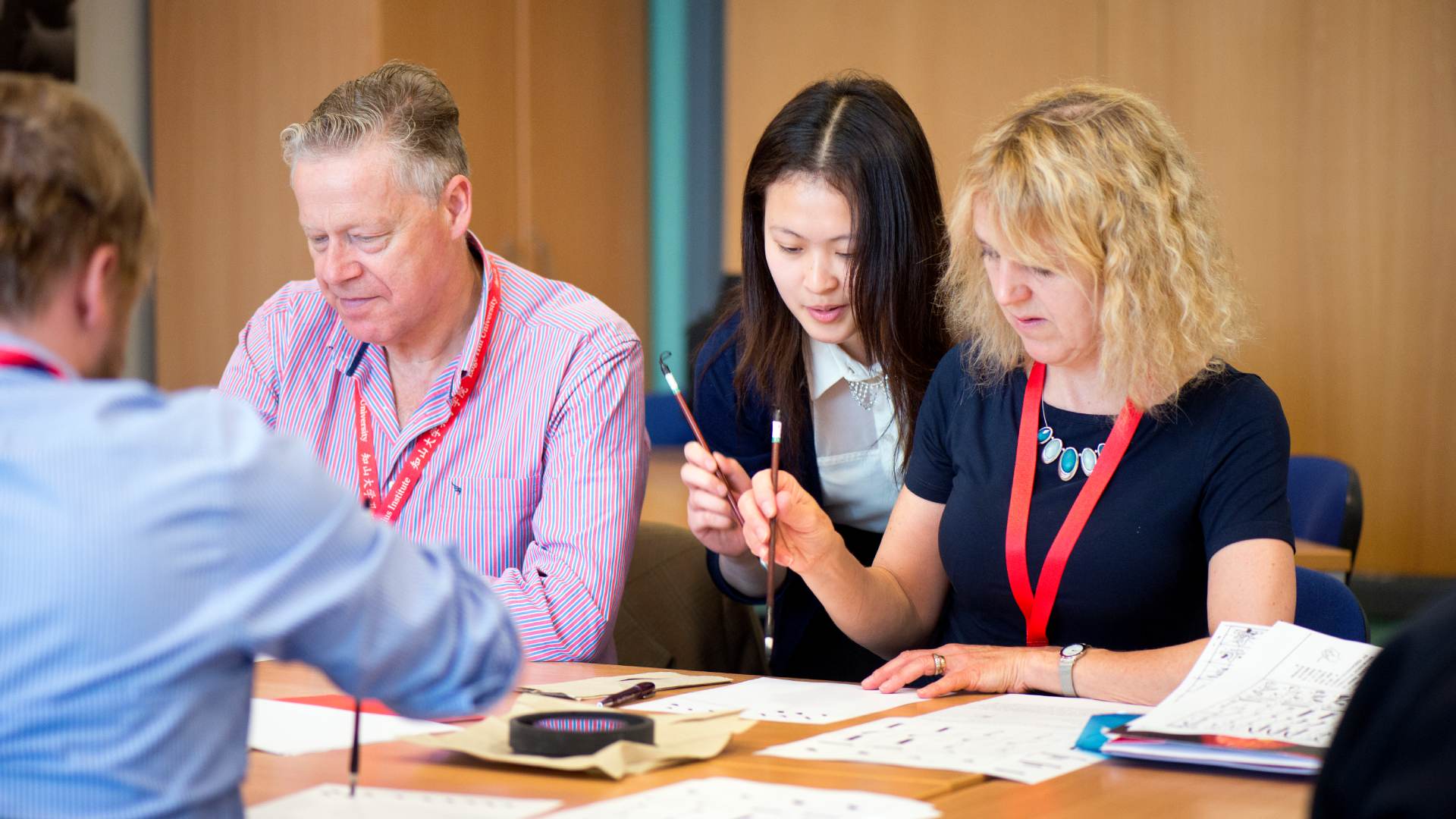 A picture of two students sat at a desk, with a Mandarin Lecturer sat with them, helping them with their Mandrian writing.