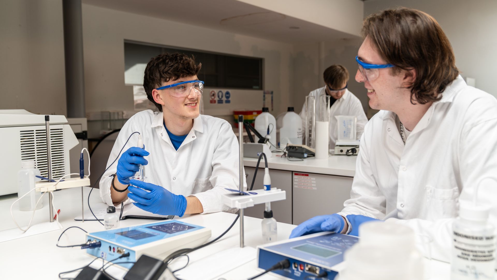 Two students wearing white coats and blue gloves work in a lab.