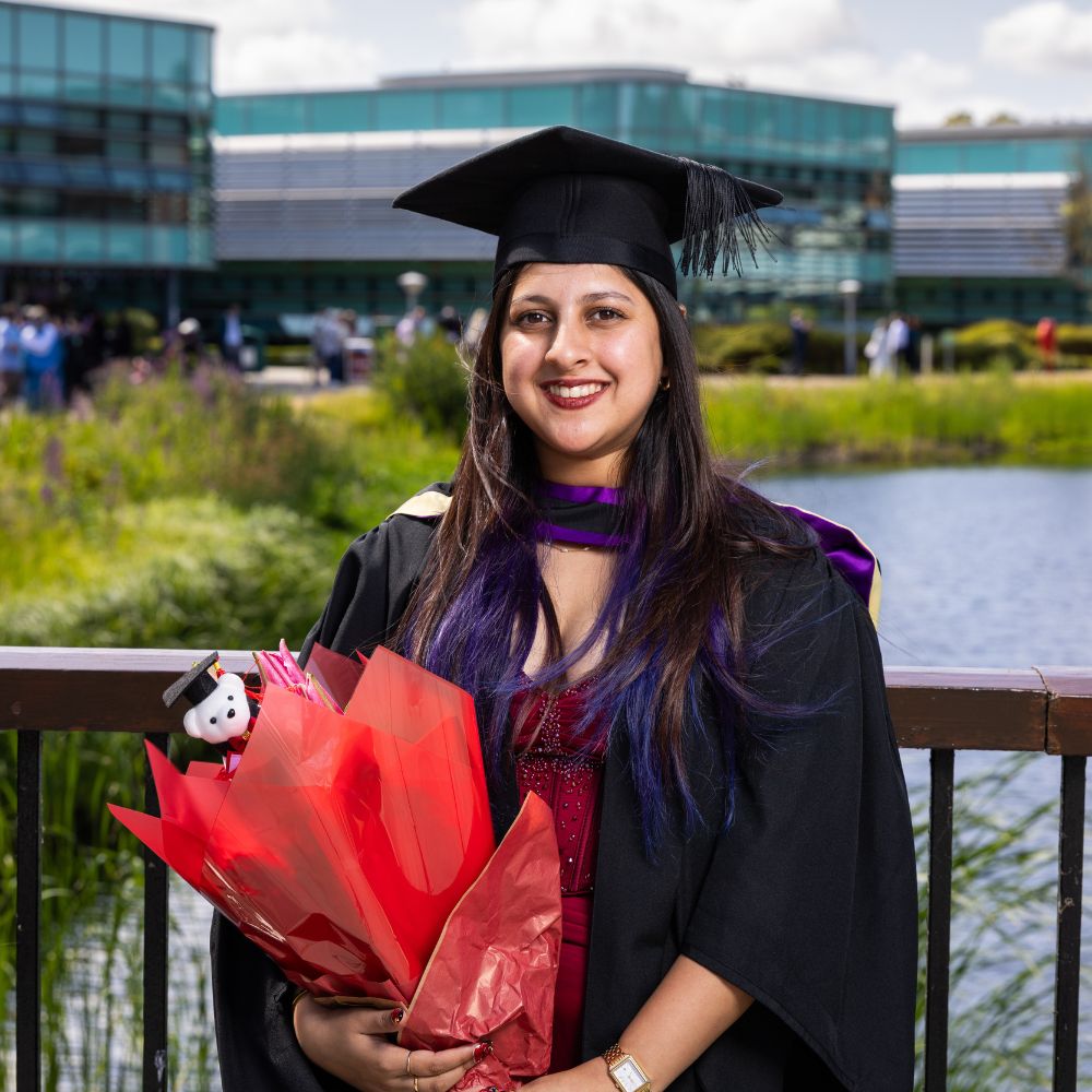 Isha Bhukya at graduation in their gown and hat with a bouquet of flowers in hand.