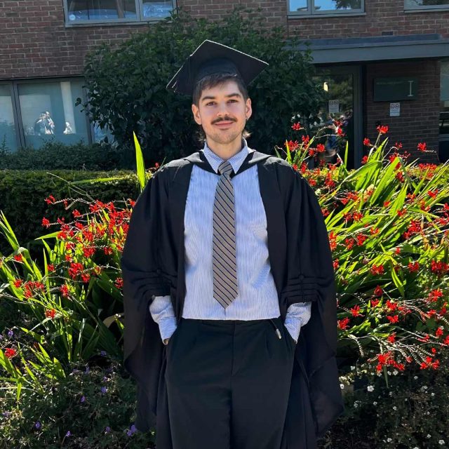 Jarod Purvis in their cap and gown on graduation day, standing in front of orange flowers on campus