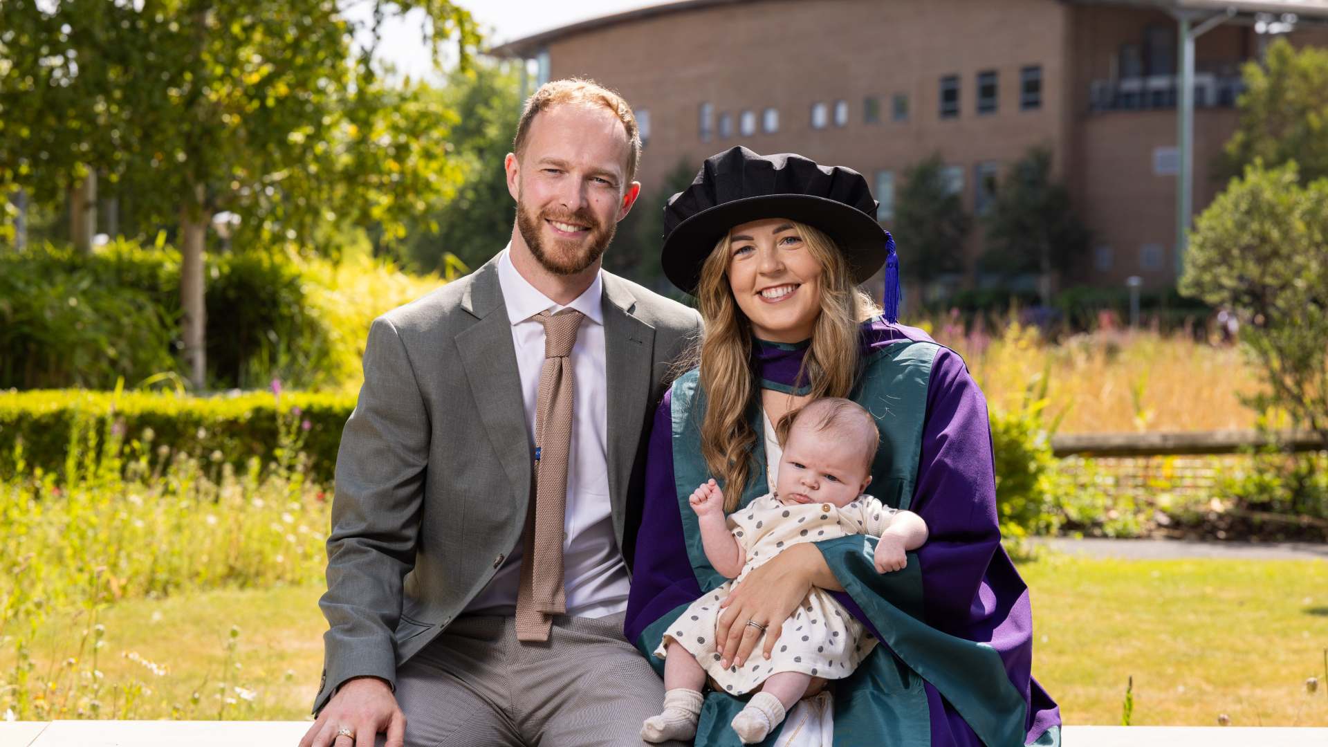 Simone Magill wears a cap and gown, holding her baby Skye and sitting next to her husband Mark.