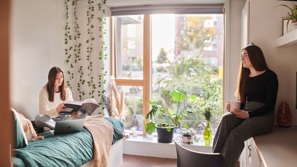 Two students in a bedroom in one of our our accommodation blocks. One is sat on the bed with their laptop reading from a textbook, and the is sat with a mug.