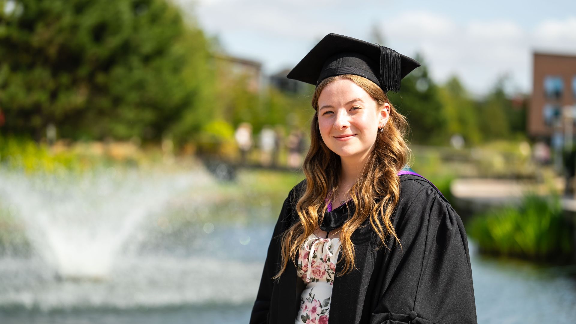 student with graduation outfit and hat with water behind her.