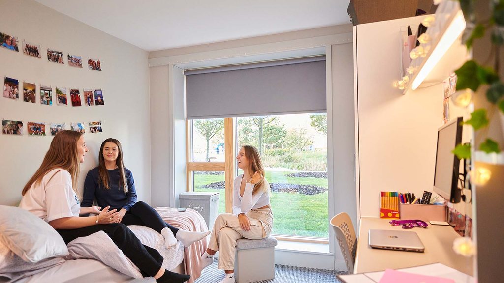 Students sat in a room in Chancellors South. Two are sat on a bed and one on a stool next to them. There are photos hung on the wall behind them.