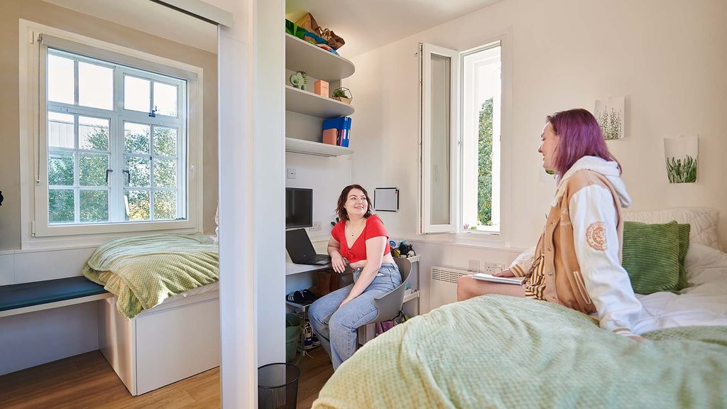 Two students sat in Main Halls accommodation. One is sat on a bed and the other sat on a desk with a laptop and shelves with folders on.